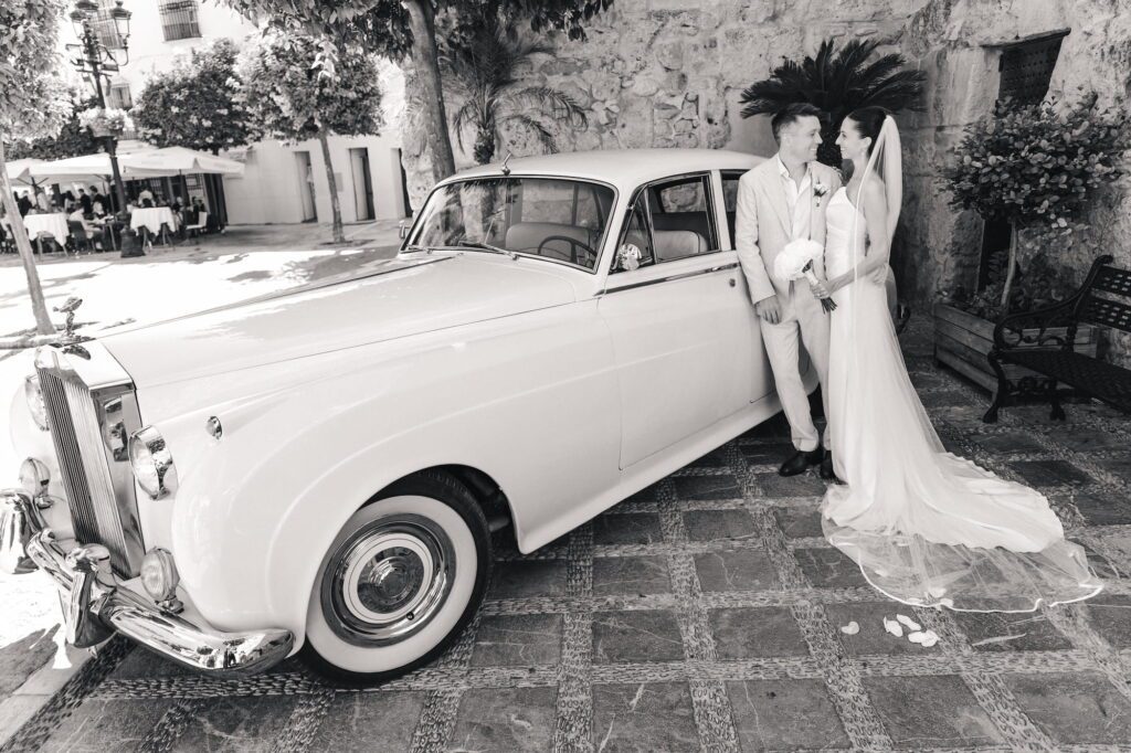 Anna and Michael standing face to face beside a Rolls-Royce in a Marbella courtyard, photographed in black and white