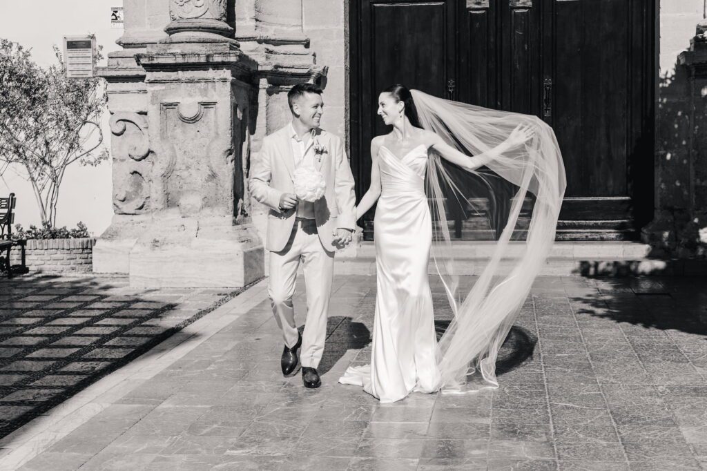 Anna and Michael walking outside the church as Anna spreads her veil in the wind, photographed in black and white