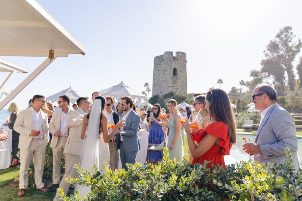 Wedding guests holding Aperol Spritz cocktails outside a bar beside the pool