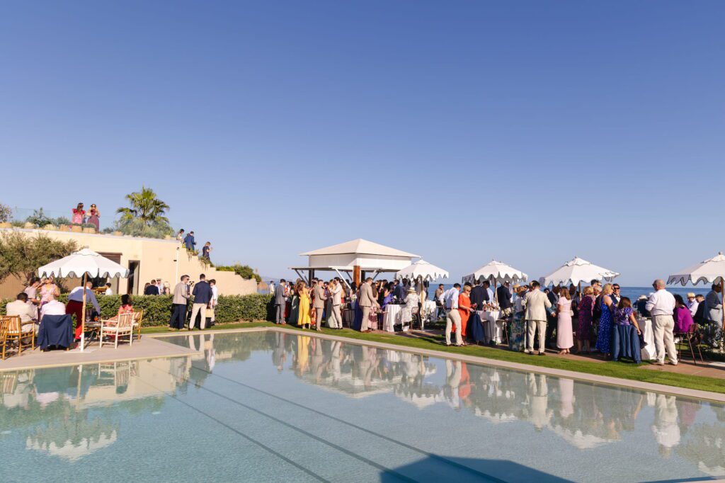 Wide view of wedding guests gathered around the poolside area beneath parasols
