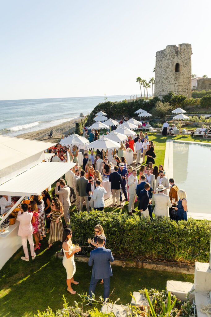 Wedding guests gathered at the poolside bar with the Mediterranean coast and a distant castle ruin visible beyond