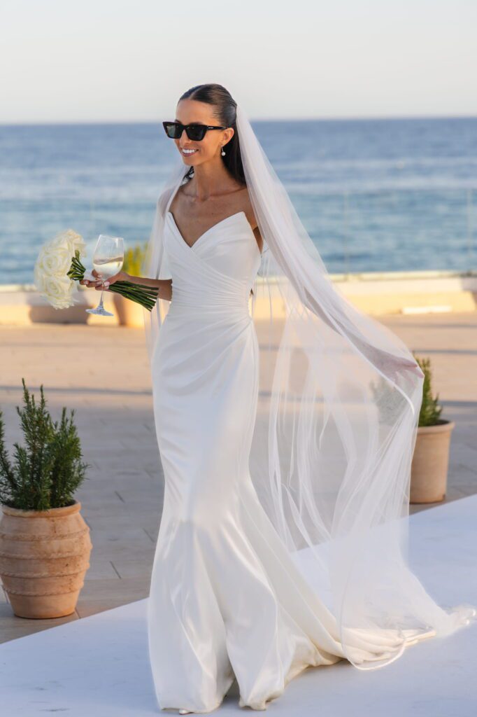 Anna walking through the wedding venue carrying a glass and bouquet while wearing sunglasses
