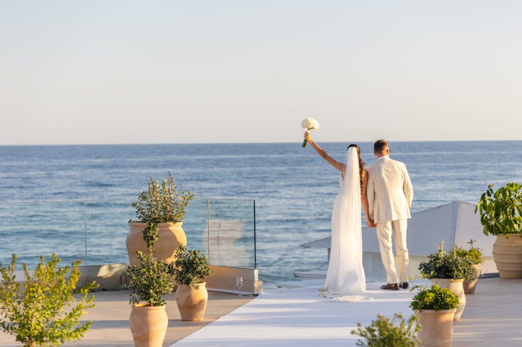 Anna and Michael standing hand in hand facing the ocean, with Michael leaning towards Anna as she raises her bouquet